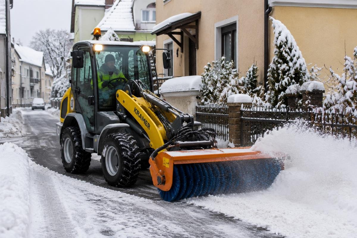 A compact wheel loader with a snow brush attachment clearing a narrow residential street during winter